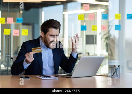 Frustrated businessman with credit card having problems with online transaction in office. Man in business suit showing anger while looking at laptop screen using credit card for e-commerce. Stock Photo