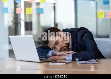 Exhausted businessman in suit sleeping on desk beside laptop and documents in modern office. Concept of workplace fatigue, overwork, stress, and work-life balance among office workers. Stock Photo