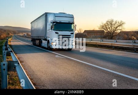 Big white semi trailer truck on a highway driving at bright sunny sunset. Transportation vehicle Stock Photo