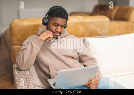African american man in brown hoodie using laptop, studying, learning, shopping, working online,   while sitting on a sofa at home Stock Photo