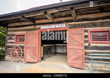 Valley Tools And Trades Log Barn Exterior Shenandoah Heritage Village Luray Virginia | LURAY, Virginia, United States — Red plank doors open beneath a hand-lettered "Valley Tools & Trades" sign on a log exhibit building at Shenandoah Heritage Village, the seven-acre open-air museum on the Luray Caverns grounds. The structure is one of several endangered Shenandoah Valley buildings rescued and rebuilt here from salvaged logs, stone, and timber. A weathered wagon wheel leans against the chinked log wall, marking the agrarian theme of the collection inside. Stock Photo