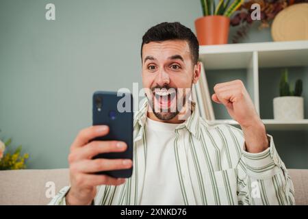 Young man celebrating that he has won a sports bet while checking the result on a smartphone app. A happy guy betting at an online casino with a cellphone. An excited male watching the lottery prize. High quality photo Stock Photo