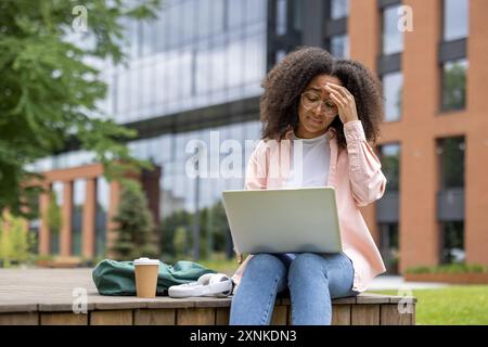 Stressed african american woman student working on laptop on campus. She appears frustrated and exhausted, sitting outside university building with coffee and backpack, showing fatigue with studies. Stock Photo