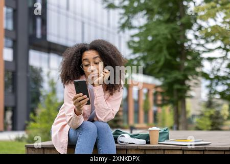 African American woman student sitting outdoors using smartphone, showing bored expression. Surrounded by laptop, coffee cup, and headphones. Modern urban background with greenery and buildings. Stock Photo