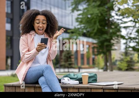 Happy African American woman student using smartphone with joyful expression outdoors on campus. Wearing glasses and casual attire. Open laptop, coffee cup, and headphones on the table. Stock Photo