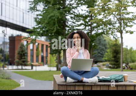 Happy African American female student sitting on campus bench with laptop, enjoying outdoor study session. Casual attire, backpack, coffee, and relaxed vibe reflect college life academic dedication. Stock Photo