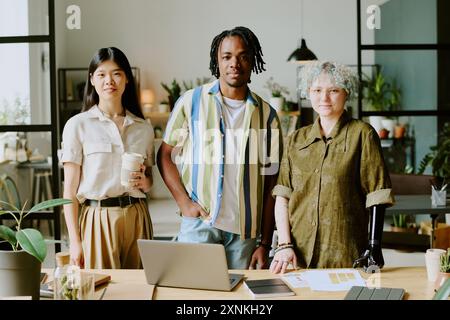 Medium long shot of three diverse gen Z coworkers posing for camera at work in modern eco-friendly office Stock Photo