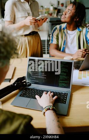Vertical over shoulder shot of unrecognizable female programmer with artificial arm coding on laptop while her colleagues having conversation Stock Photo