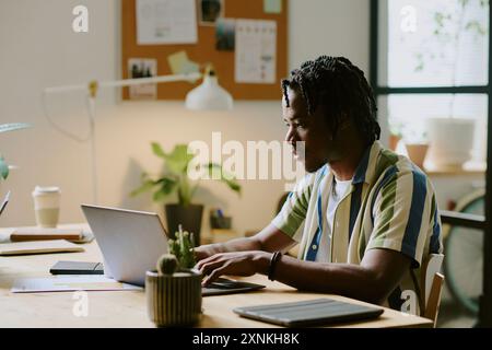 Young African American man wearing stylish casual clothes typing on laptop while sitting in modern eco-friendly office Stock Photo