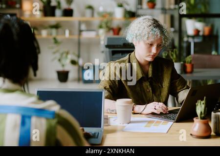Over shoulder shot of gen Z female developer wearing eyeglasses sitting in front of her colleague working on laptop Stock Photo