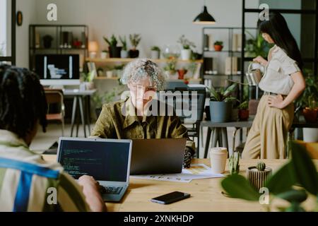 Diverse young programmers developing codes on laptops while their female colleague watering plants in modern biophilic office Stock Photo