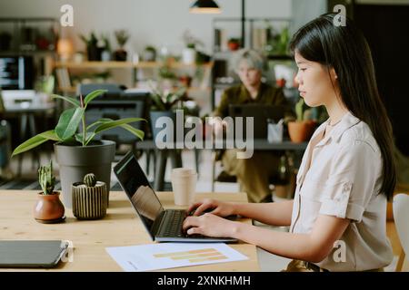 Side view of young Asian woman wearing casual clothes typing code on laptop at workplace in modern eco-friendly office with biophilic design Stock Photo