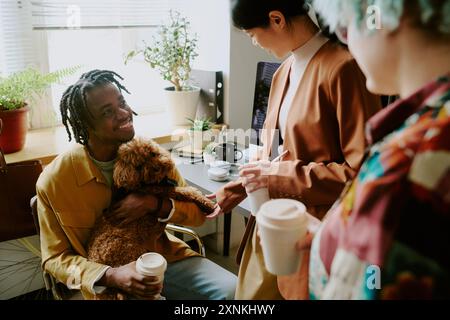 Cheerful young Black man introducing his lovely dog to female colleagues at working in modern pet-friendly office Stock Photo