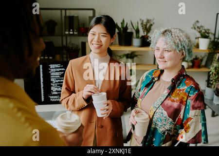 Group of three diverse gen Z colleagues holding reusable cups having conversation while standing in office during coffee break Stock Photo