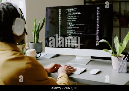 Over shoulder shot of unrecognizable young African American programmer wearing headphones coding on modern desktop computer in office Stock Photo