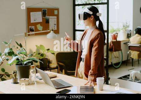 Young Asian female programmer wearing VR headset testing new app at work in modern IT company office, copy space Stock Photo