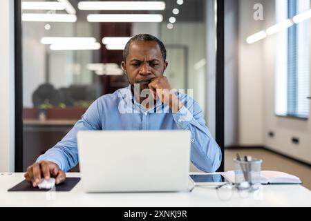 Mature african american office worker deeply focused at desk inside modern workspace. Displaying concentration and dedication while working on laptop in professional office environment. Stock Photo