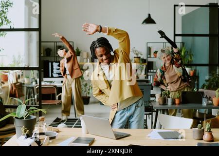 Group of cheerful young ethnically diverse coworkers doing morning exercise in modern office with biophilic interior design Stock Photo