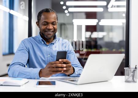 Mature african american office worker using smartphone modern workplace. Businessman interacting with mobile device, smiling during work. Technology facilitating professional communication in office Stock Photo