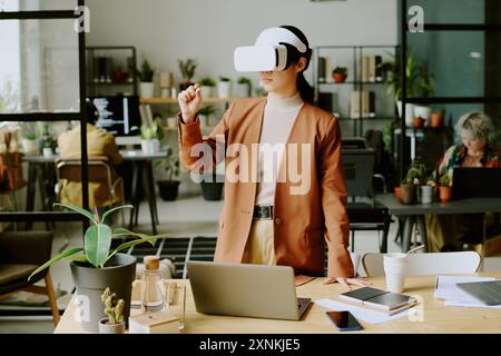 Young Asian female developer wearing headset using VR software at work in modern eco-friendly office of IT company Stock Photo