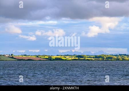 Gentle waves lap against a vibrant shoreline, as rolling hills bask in soft light beneath a brooding sky at dusk. Stock Photo