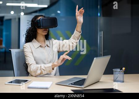 Businesswoman using virtual reality headset in modern office, interacting with digital interface. Young woman immersed in VR technology, exploring virtual workspace, innovation in business Stock Photo