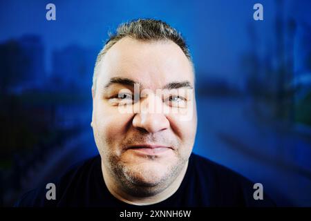 Photographing face with wide-angle lens from close range, 47 year old Caucasian man with an excess weight problem, portrait on blue background. Stock Photo