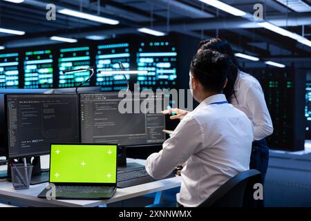 Team of technicians in data center using isolated screen laptop to find firewall misconfigurations affecting servers. IT staff members in server room checking systems bottlenecks using mockup device Stock Photo