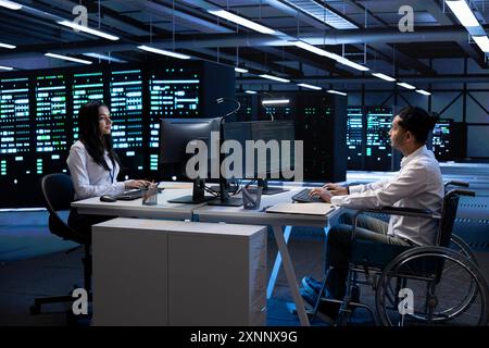 Person with paraplegia and colleague working in inclusive server hub workspace, typing on PC. Man in wheelchair and coworker doing tasks in data center housing advanced data storage infrastructure Stock Photo