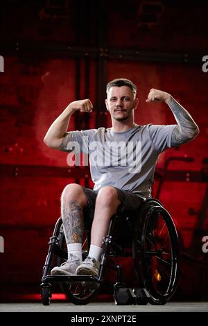Motivational full length portrait of tattooed young man using wheelchair in gym and flexing arm muscles on red background Stock Photo
