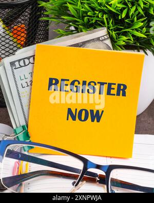 REGISTER NOW words written on a yellow sticker on the table Stock Photo