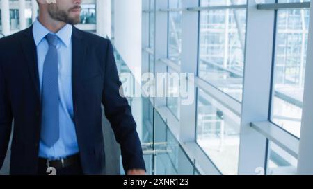 An American businessman walks inside a modern building while leaning on a glass window inside the building. Confident young businessman lost in though Stock Photo