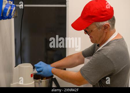 A man wearing a red cap and blue gloves is mixing ingredients in a kitchen. He is focused on his task, using a mixer. The kitchen has a clean, modern Stock Photo