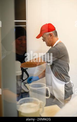A man in a red cap and apron is using a mixer in a kitchen. He is focused on his task, with blue gloves on, preparing food. A reflection of another pe Stock Photo