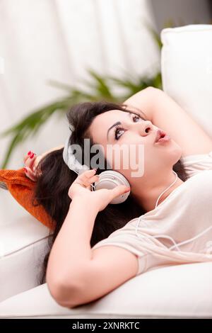 Young woman lies on her sofa at home, listening to music with headphones, feeling happy and relaxed in her modern living room Stock Photo