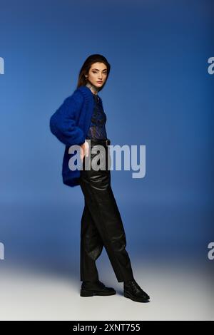A young woman in a blue cardigan and black leather pants poses against a blue background. Stock Photo