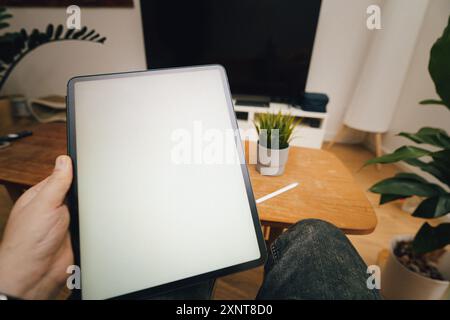 First person view. Hand holding tablet. Empty white screen. Home interior. Stock Photo