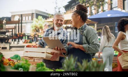 International Adult Partners Working at a Farmers Market. Diverse Couple Selling Ecological Fruits and Vegetables From an Outdoors Food Stand. Adult Female Using Laptop to Communicate with Suppliers Stock Photo