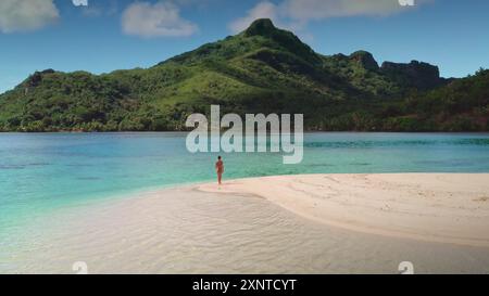 Woman wearing a bikini is enjoying a peaceful walk on the beach of a tropical island with lush green mountains in the background Stock Photo