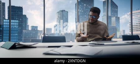 Stressed Out Balck Businessman at a Desk in Skyscraper Office, Using Laptop Computer. Tired Manager Completing Complicated Financial Report Before Deadline, Feeling Overwhelmed. Stock Photo