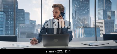 Confident Businessman in a Suit Sitting at a Desk in Office, Using Laptop Computer and Picking Up A Call On Smartphone From Colleague. Successful Sales Manager Discussing Project. Stock Photo