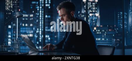 Successful Handsome Businessman Working on Laptop Computer in Big City Office Late At Night. Financial Investment Analyst Checking Data And Works On Risk Management In Hedge Fund. Stock Photo