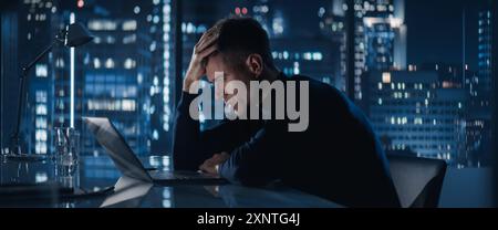 Distraught Businessman Working on Laptop Computer in Big City Office Late At Night. Financial Investment Analyst Checking Data And Feeling Stressed Due To Negative Corporate Report. Stock Photo