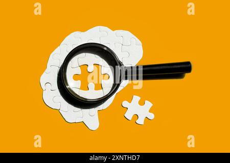 Brain made of white puzzle pieces, with a magnifying glass focusing on an empty puzzle piece isolated on yellow backdrop. Mental health, cognition, an Stock Photo