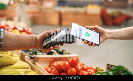 Close Up of a Customer Making a Secure Contactless Online Payment Using a Smartphone with a Credit Card Banking NFC App. Modern Shopper Buying Organic Fruits and Vegetables From a Farmers Market Stall Stock Photo