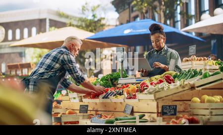 Multiracial Adult Partners Working at a Farmers Market. Diverse Couple Selling Ecological Fruits and Vegetables From an Outdoors Food Stand. African Vendor Using Laptop to Communicate with Suppliers Stock Photo
