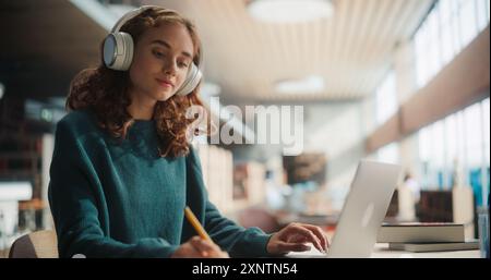 Engaged Young Caucasian Female Student Studying With Headphones in Modern Library. Woman Using Laptop and Taking Notes, Focused on Academic Research and Learning in a Public Educational Environment Stock Photo