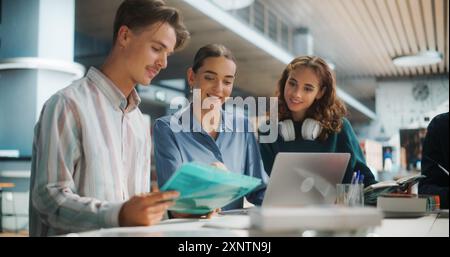 Engaged Group of University Students Collaborating on a Project in a Modern Library. Two Women and One Man Discussing Academic Material Over a Laptop and Books, Focused and Smiling. Stock Photo