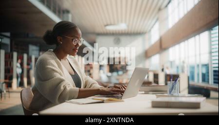 Dedicated African American Female Student Engaged in Online Learning at a Modern Library. Young Woman Using Laptop for Research, Surrounded by Books and Academic Environment. Stock Photo