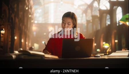 Diligent Asian Female Student Engaged in E-Learning with a Laptop and Books in a Classical Library. Young Woman Focused on Studying for University Exams, Surrounded by Historical Architecture. Stock Photo